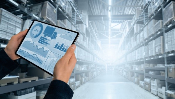 two hands holding a tablet showing an on-screen analytics platform that's connected to the stock in the background warehouse setting. 