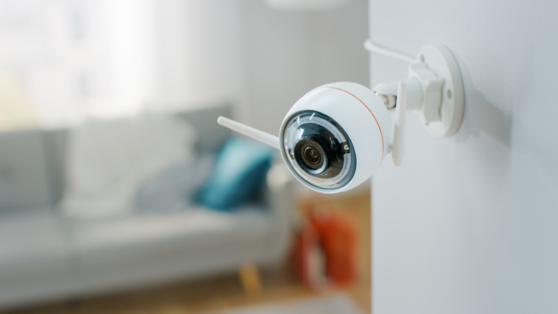 Modern Wi-Fi Surveillance Camera with Two Antennas on a White Wall in a Cozy Apartment. Man is Sitting on a Sofa in the Background.