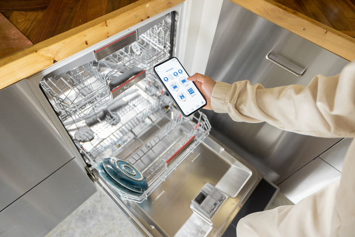 Woman holding phone with running smart home application, controlling dishwasher remotely. Smart kitchen appliances concept.