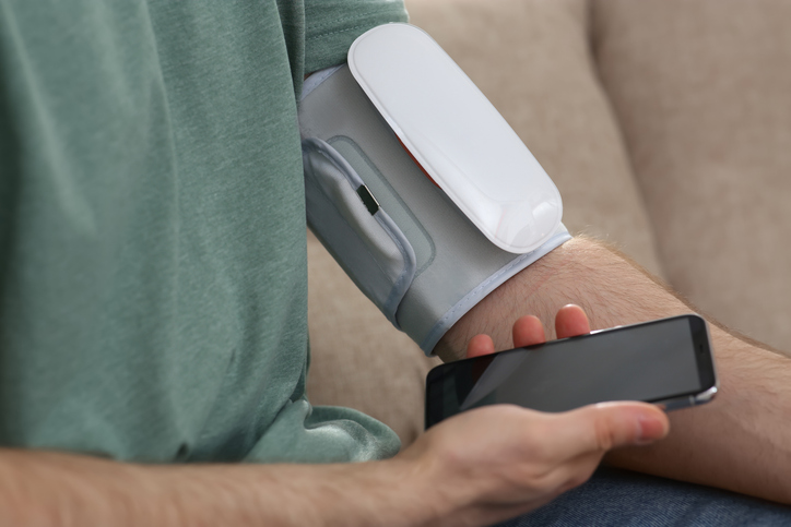 Man checking blood pressure with modern monitor and smartphone indoors, closeup.