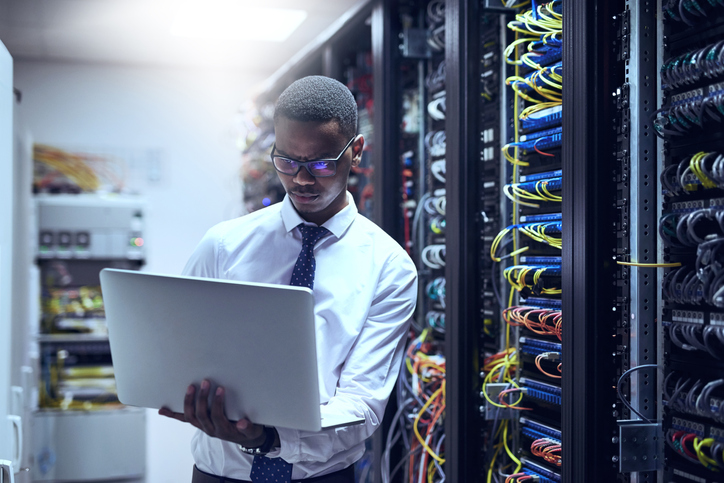 IT technician working on his laptop while standing inside of a server room.