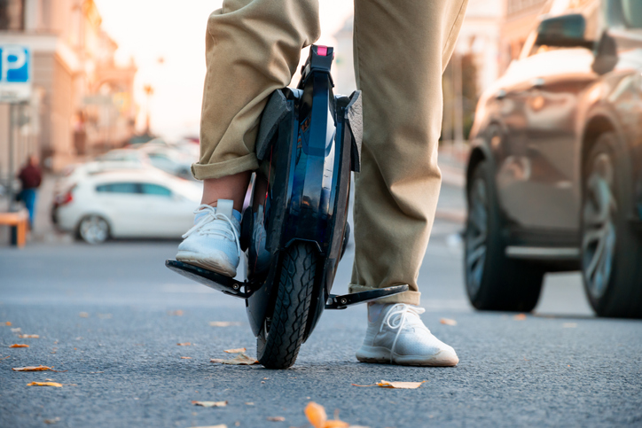 The legs of a young woman ready to ride an electric unicycle against the background of evening or morning city traffic.