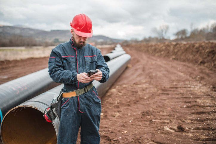 An engineer in a uniform and with a helmet on his head uses a mobile phone while controlling the operation of the gas pipeline.