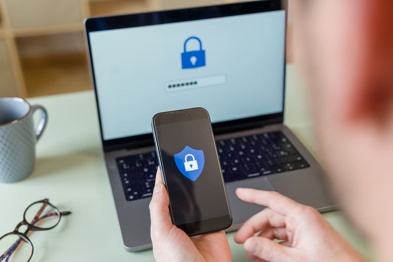 Close-up of hands holding smartphone with security app open, laptop with login screen in background.